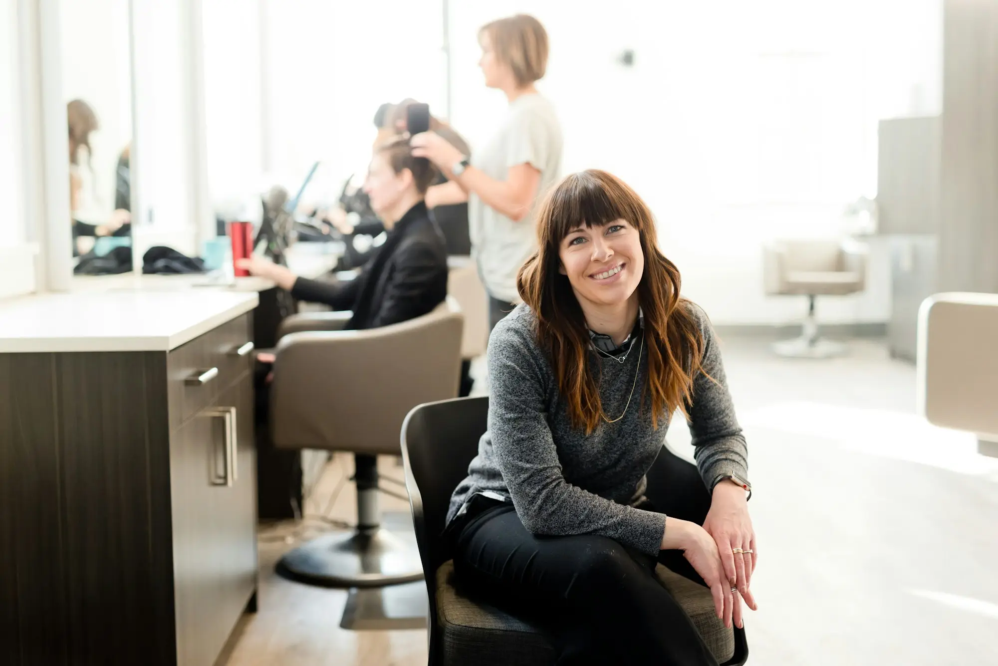 Hair salon owner sitting in a styling chair in her salon while staff work with clients behind her